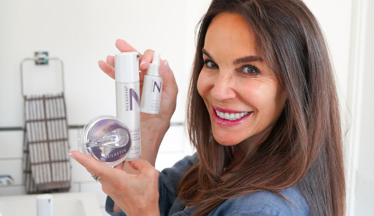 Photo of Leah Garcia,  founder and CEO of NULASTIN® looking over her  shoulder with a smile holding large and small bottles of NULASTIN®  HAIR Vibrant Scalp Treatment and a hair scrubber from her product line.