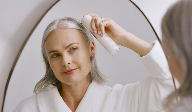Woman in a white robe applying scalp treatment from a white spray can while looking in a mirror