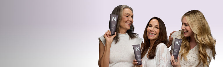 Three smiling women holding NULASTIN hair product tubes on a pale lavender gradient background