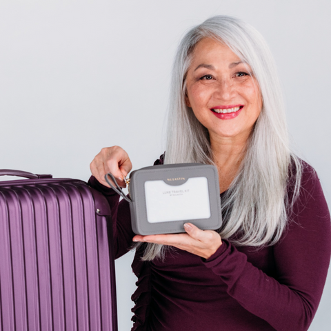 Smiling woman holding LUXE travel kit in front of purple suitcase 