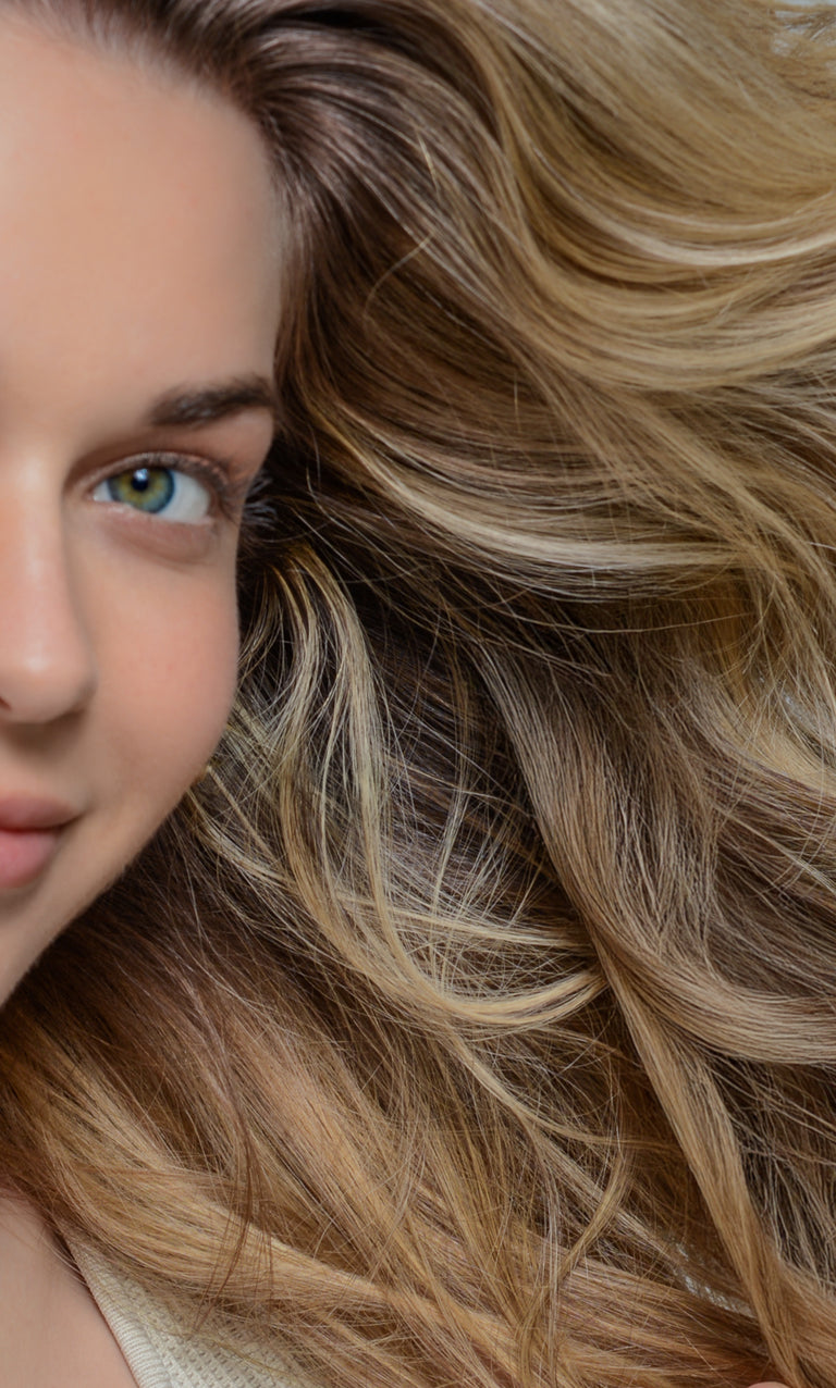 Half-face of a woman with a green eye and loose, highlighted blonde hair fanning across a light gray background