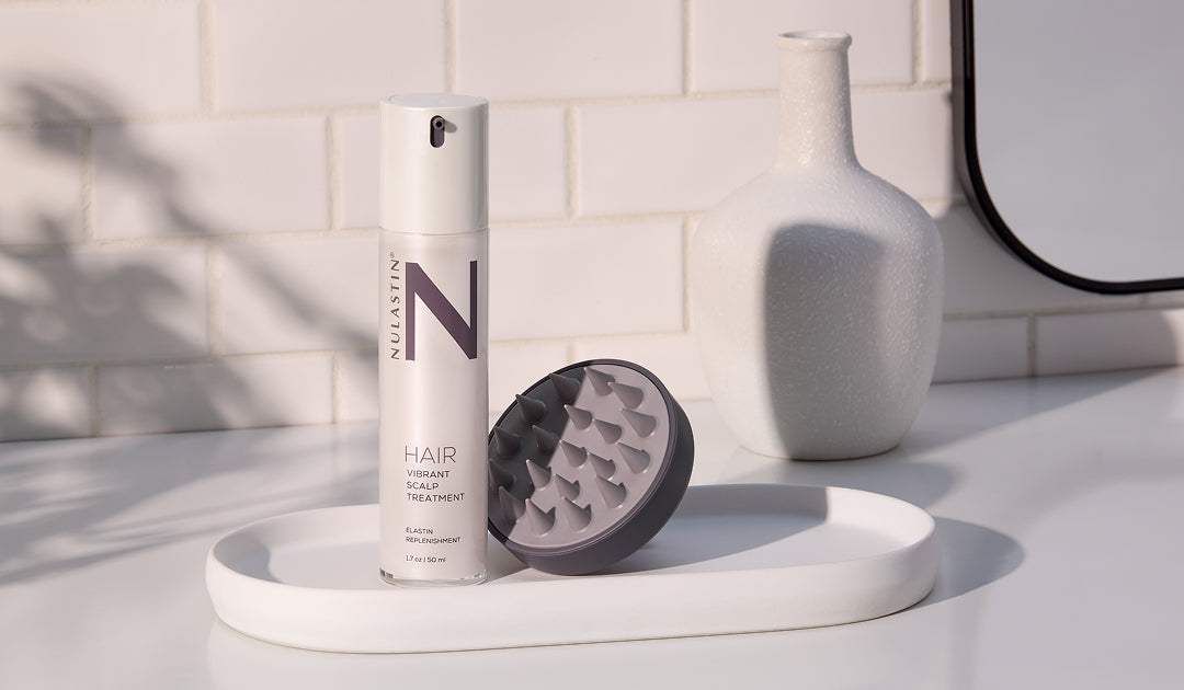 Front view of NULASTIN Vibrant Scalp Treatment bottle with silicone scalp massager on white tray in tiled bathroom