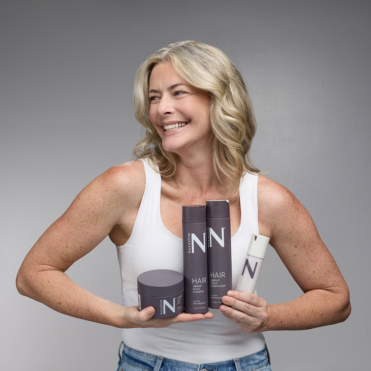 Smiling middle-aged woman holding NULASTIN hair products (shampoo, conditioner, treatment jar, serum) against a gray background