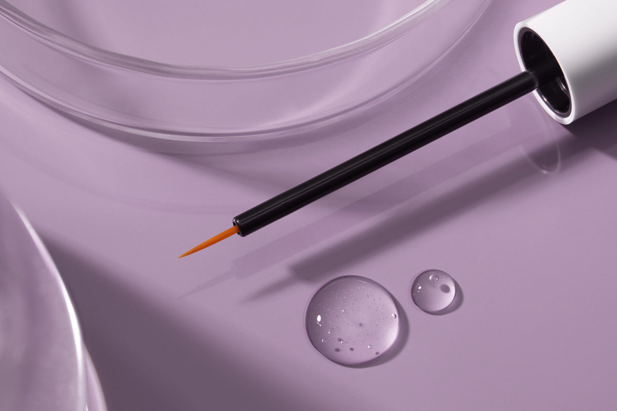 Close-up of NULASTIN LASH serum applicator with clear droplets on a lavender background