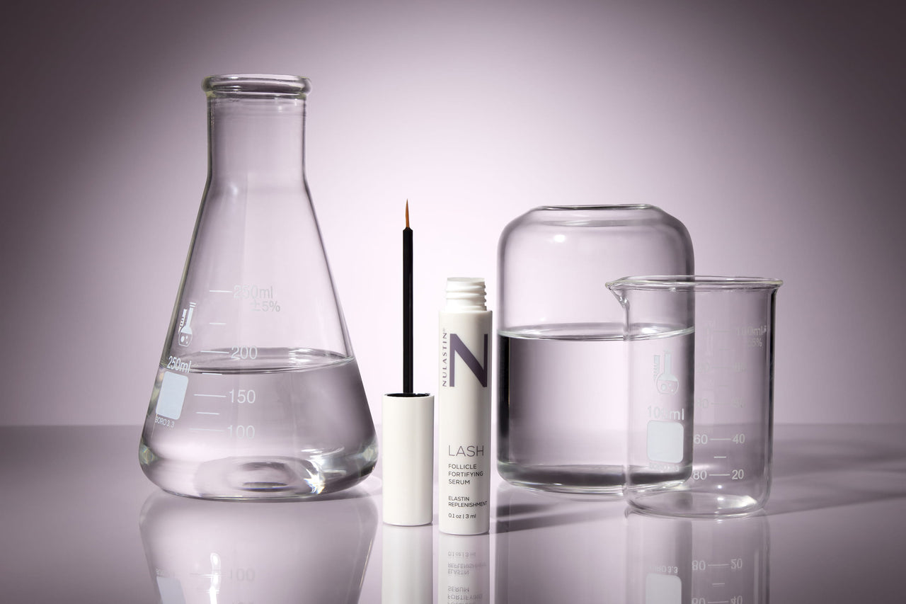 Front view of NULASTIN Lash Serum tube with applicator brush beside it, set among laboratory glassware on a reflective surface