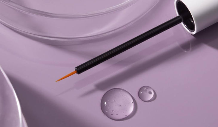 Close-up of lash serum applicator wand showing fine precision brush tip and two serum droplets on a lavender surface
