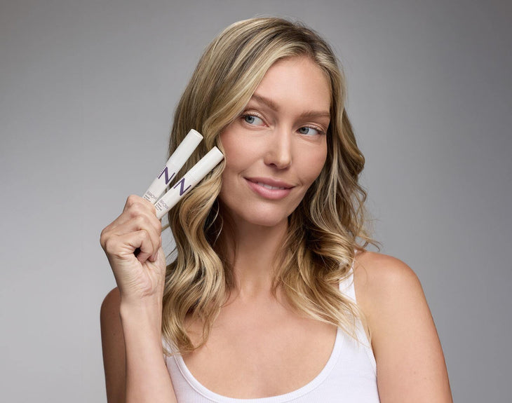 Front view of two NULASTIN Brow Serum tubes held by a model to show product size and packaging on a gray background